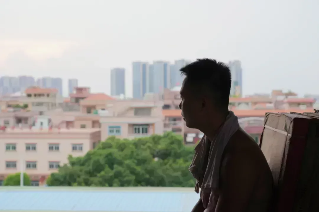 阿興在堆滿瓷磚的毛坯房休息片刻,敞開的牆體外面是一片映着落日的高樓大廈。